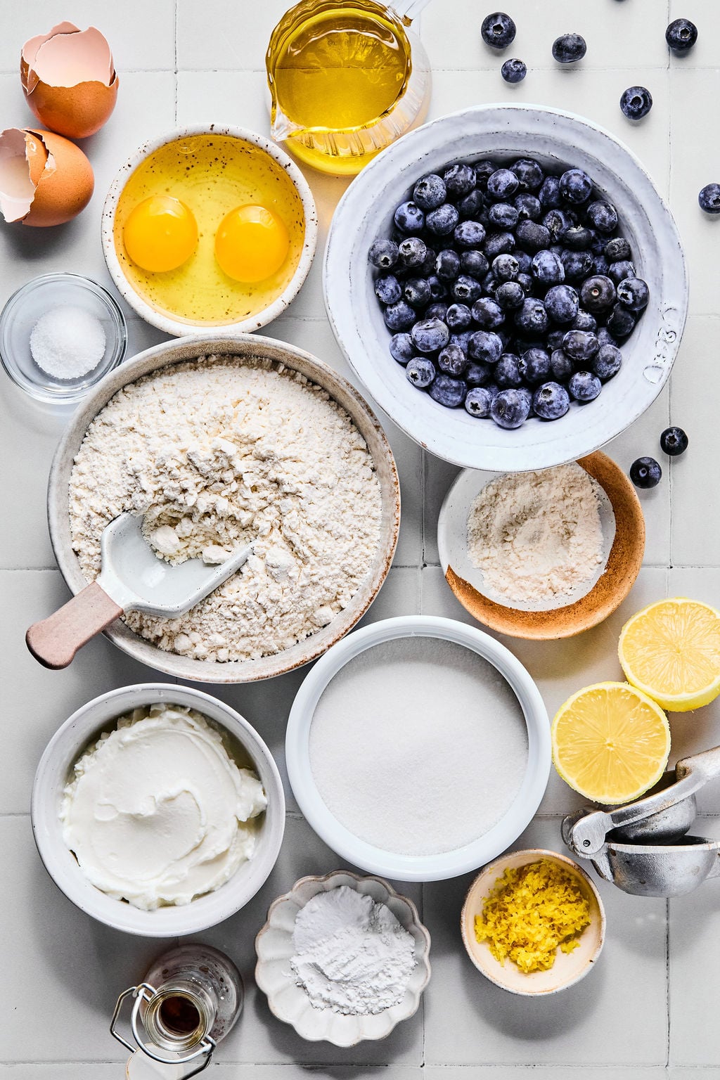 Bowls of baking ingredients including flour, sugar, eggs, blueberries, oil, yogurt, lemon, salt, baking powder, and vanilla are arranged on a white surface.