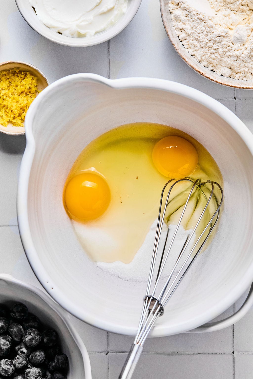 A mixing bowl with two cracked eggs, sugar, and a whisk, surrounded by bowls of flour, blueberries, lemon zest, and yogurt on a white tiled surface.