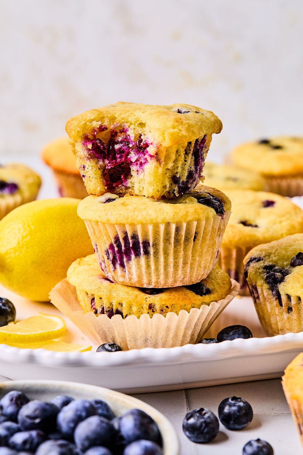 A stack of two blueberry muffins, with a bitten muffin on top, surrounded by fresh blueberries and lemons on a white plate.