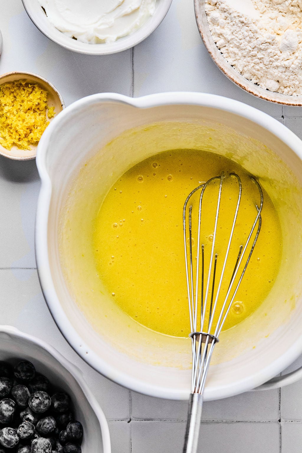A mixing bowl with a yellow batter and a whisk, surrounded by small bowls containing flour, lemon zest, blueberries, and yogurt.