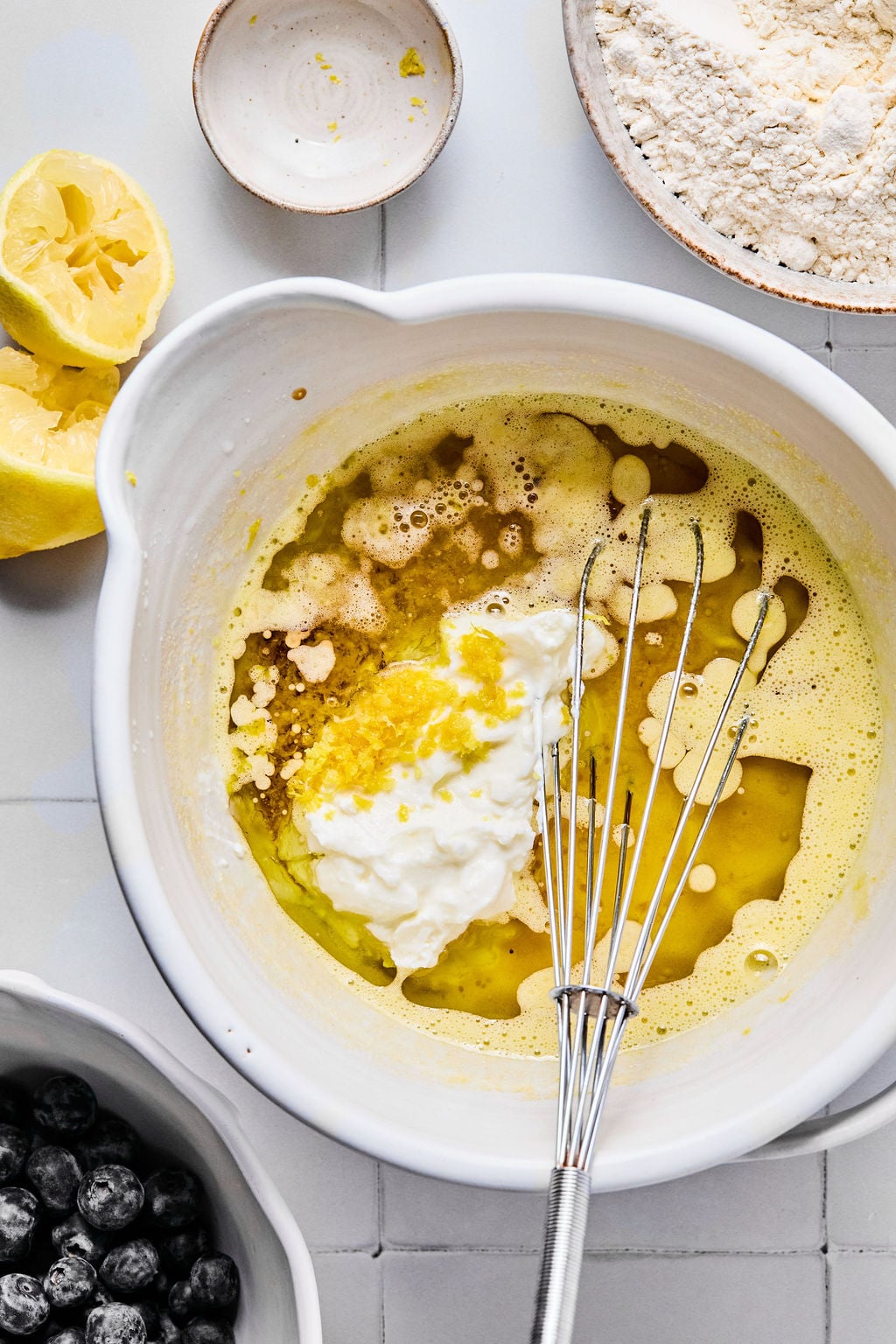 A mixing bowl with wet ingredients, lemon zest, and a whisk, surrounded by a bowl of flour, a halved lemon, and a bowl of blueberries on a kitchen counter.