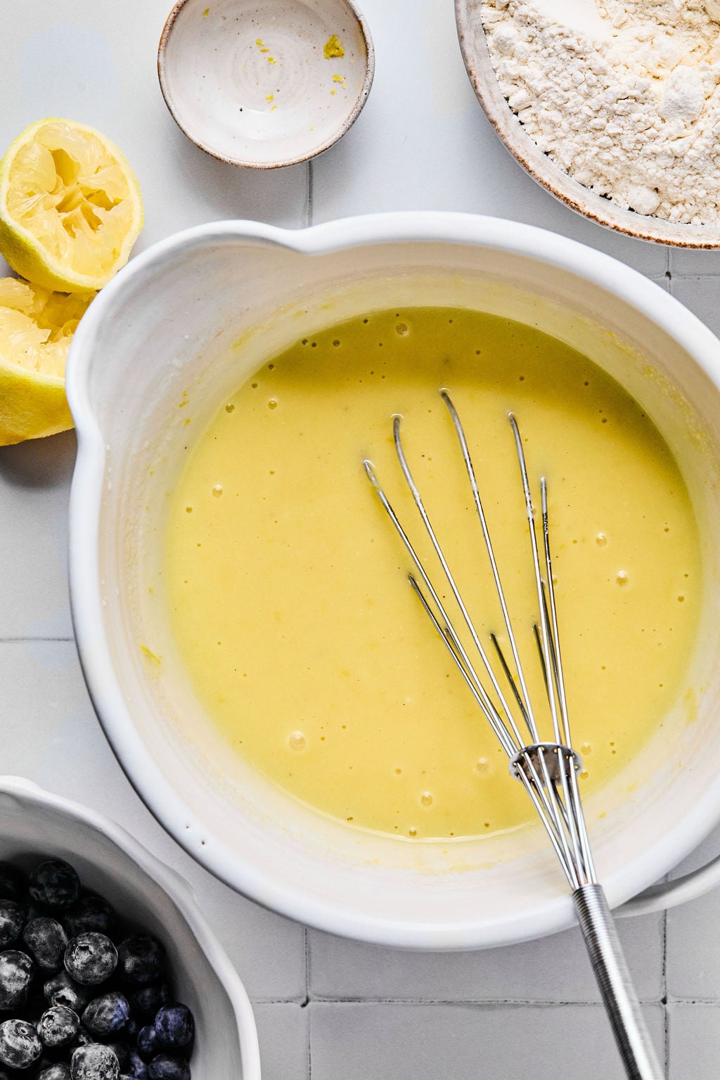 A mixing bowl with yellow batter and a whisk, surrounded by lemon halves, a bowl of flour, a small dish, and a bowl of blueberries on a tiled surface.