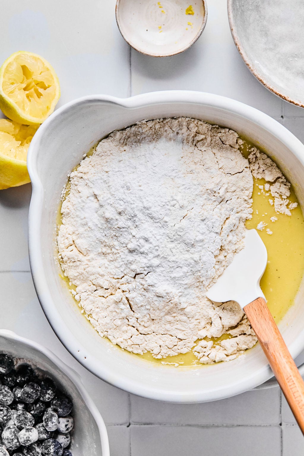 A mixing bowl with flour and wet ingredients being stirred with a spatula, surrounded by a bowl of blueberries, a squeezed lemon, and small bowls on a tiled surface.