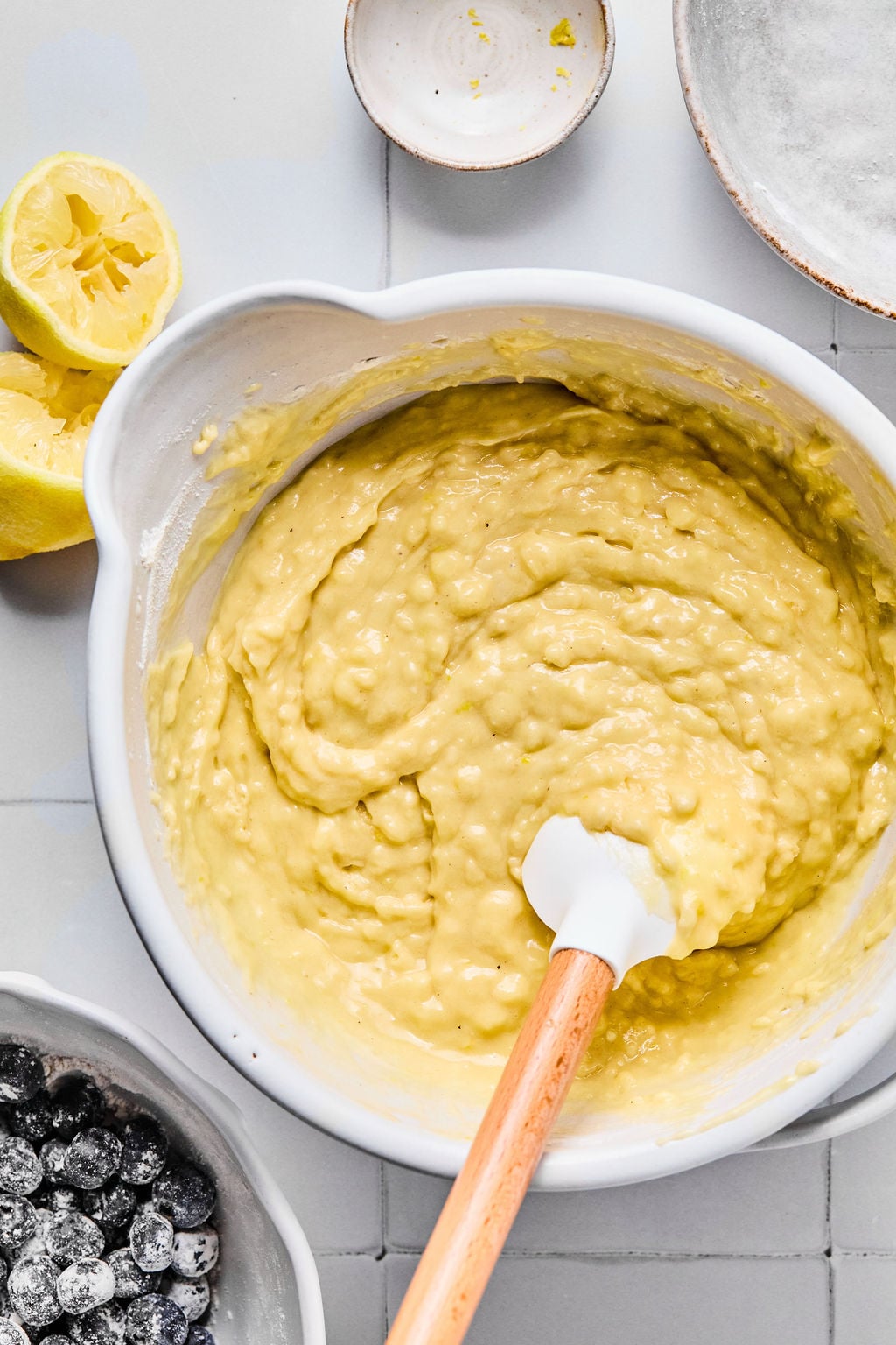 A white mixing bowl filled with yellow batter and a spatula, surrounded by halved lemons, a small dish, and a bowl of berries on a tiled surface.