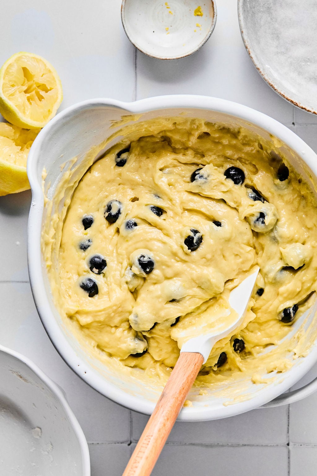 A bowl of blueberry muffin batter being mixed with a spatula, with halved lemons and empty dishes nearby on a tiled surface.