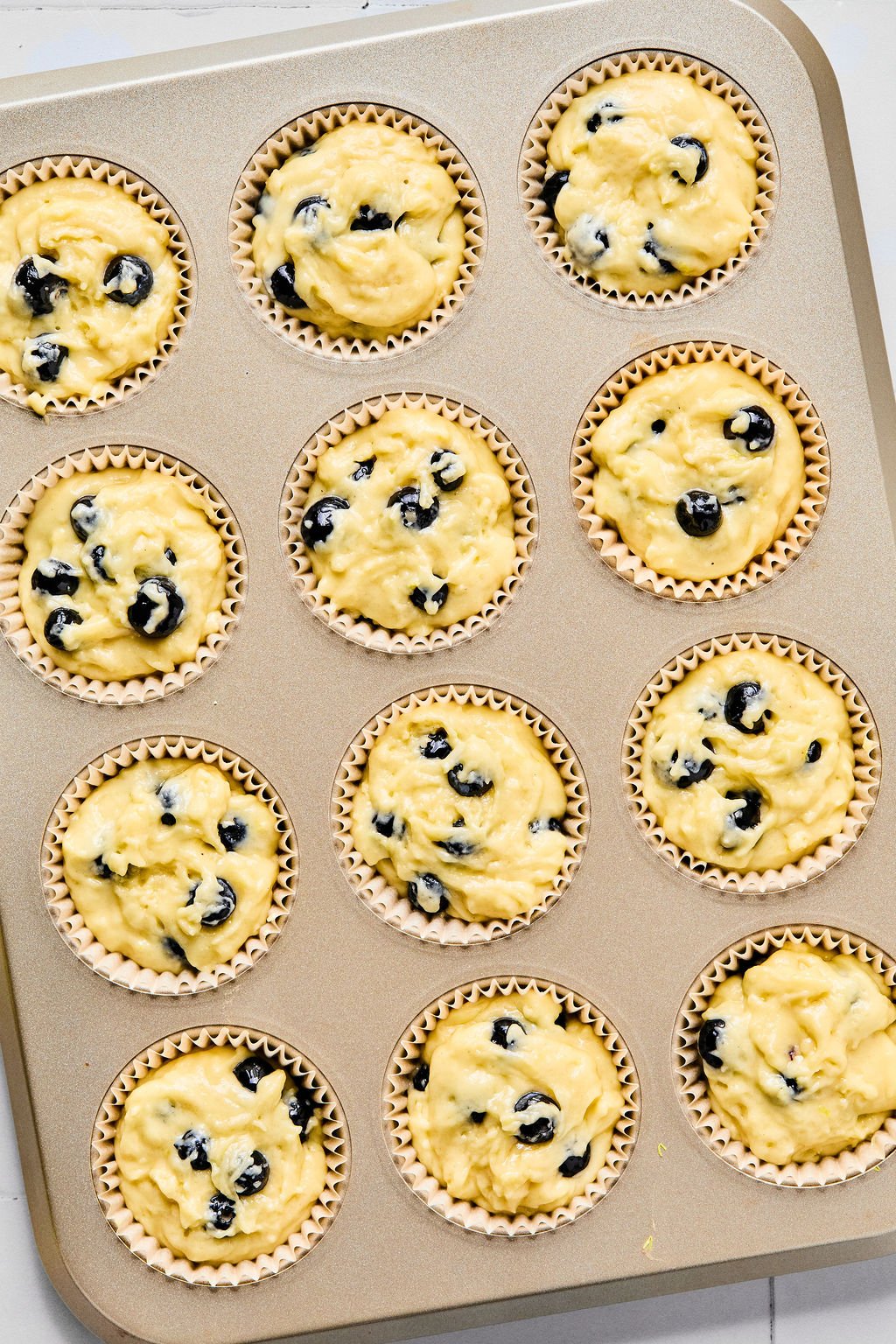 A muffin tin filled with twelve paper liners, each containing unbaked blueberry muffin batter, ready to be baked.