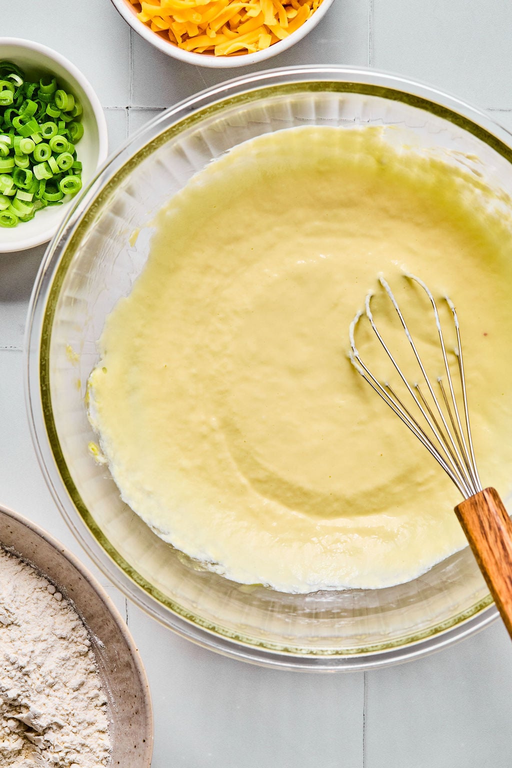 A glass bowl with a whisk mixing yellow batter, surrounded by bowls of shredded cheese, chopped green onions, and flour.