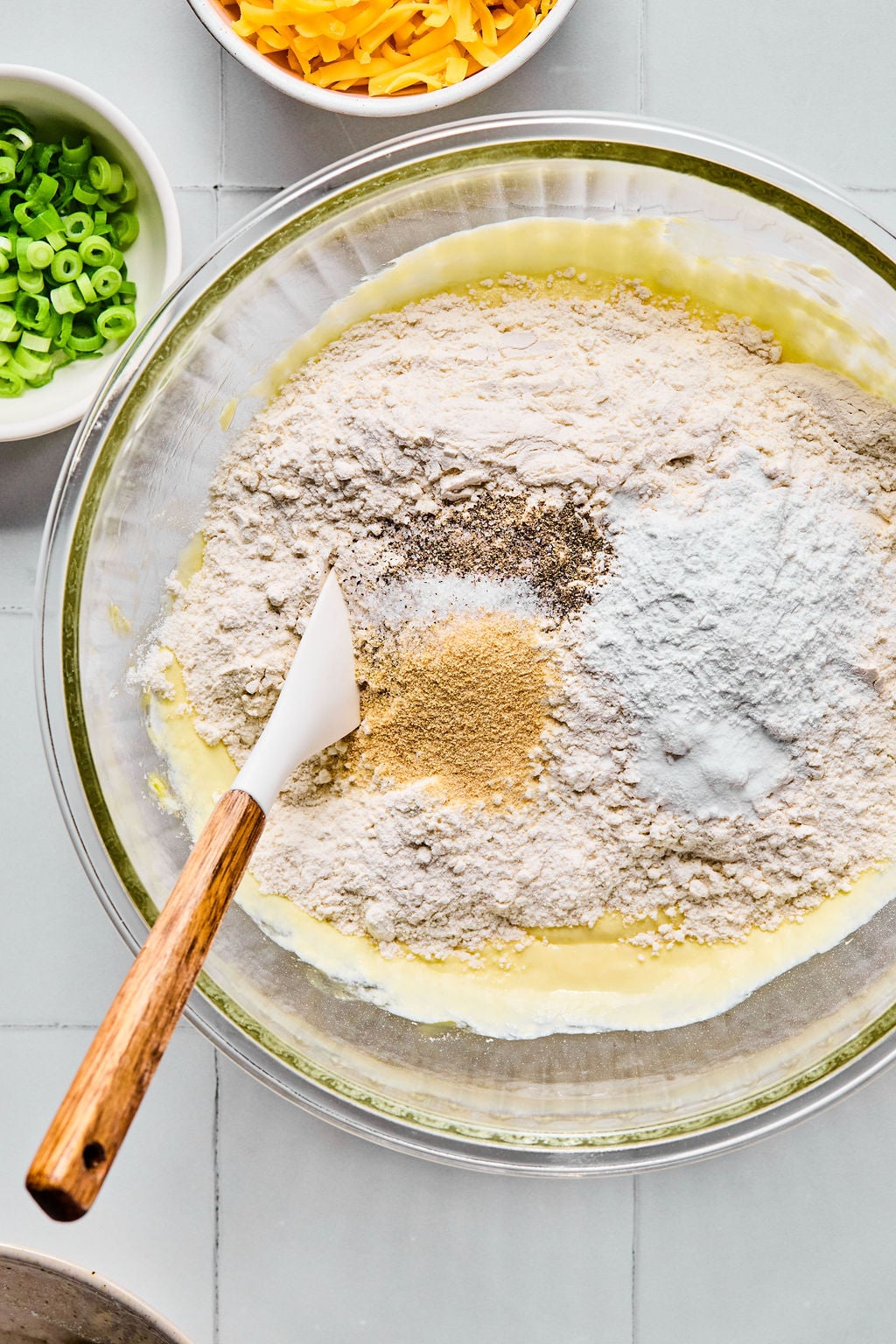 A mixing bowl with flour, baking powder, spices, and wet ingredients being combined with a spatula; bowls of green onions and shredded cheese are nearby.