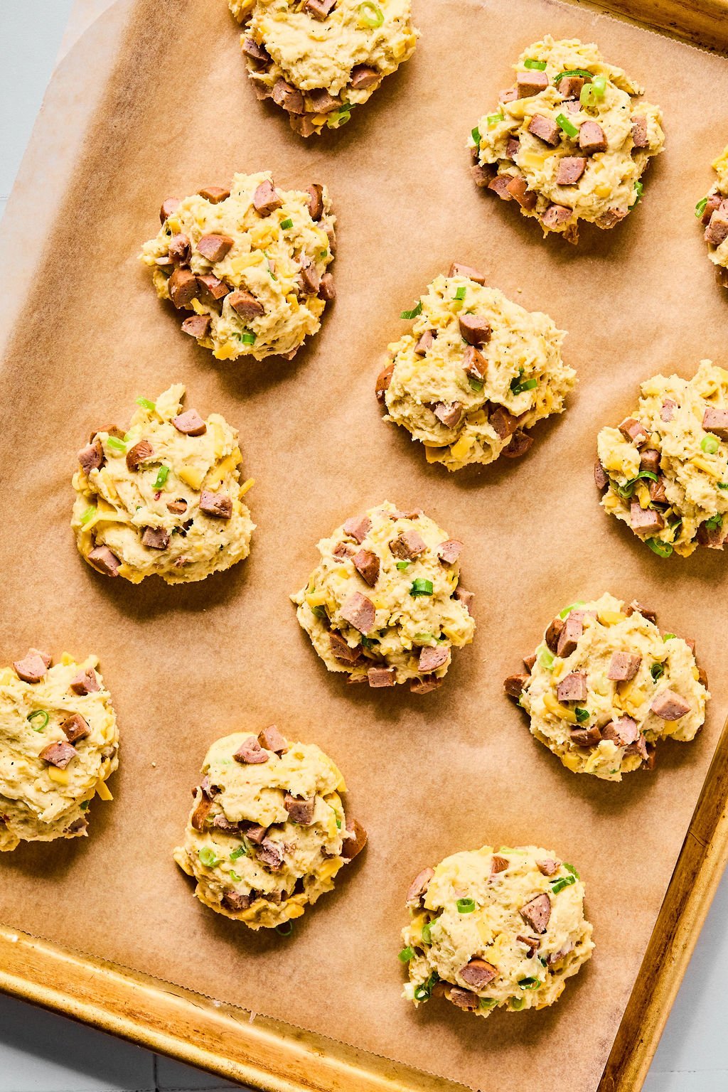 Raw, chunky dough mounds with visible chopped green herbs and sausage pieces are arranged on a parchment-lined baking sheet.