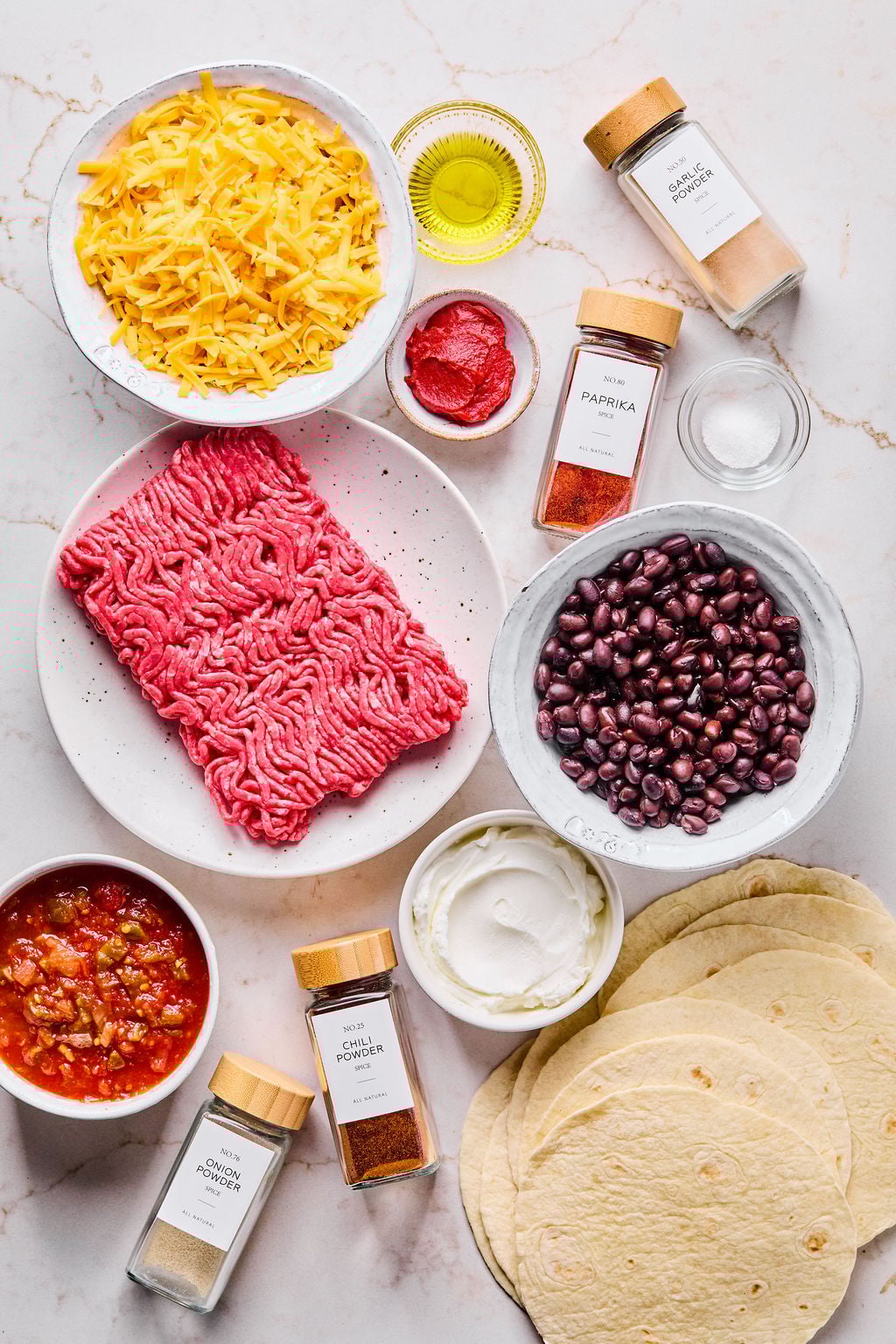 Ingredients for tacos laid out on a white surface, including ground beef, shredded cheese, black beans, flour tortillas, salsa, sour cream, tomato paste, oil, and various spices.