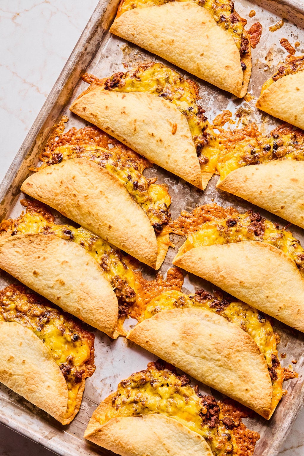 A baking sheet with several crispy taco shells filled with ground beef and melted cheddar cheese, arranged in rows.