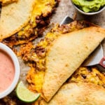 Baked quesadillas filled with melted cheese and beef, served with a lime wedge, guacamole, and a bowl of dipping sauce on the side.