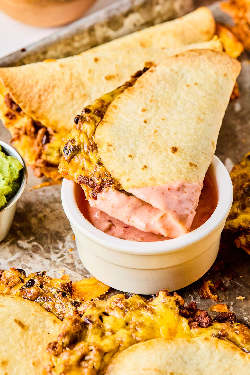 A folded taco with melted cheese and ground meat is being dipped into pink sauce; other tacos and a small bowl of guacamole are nearby.