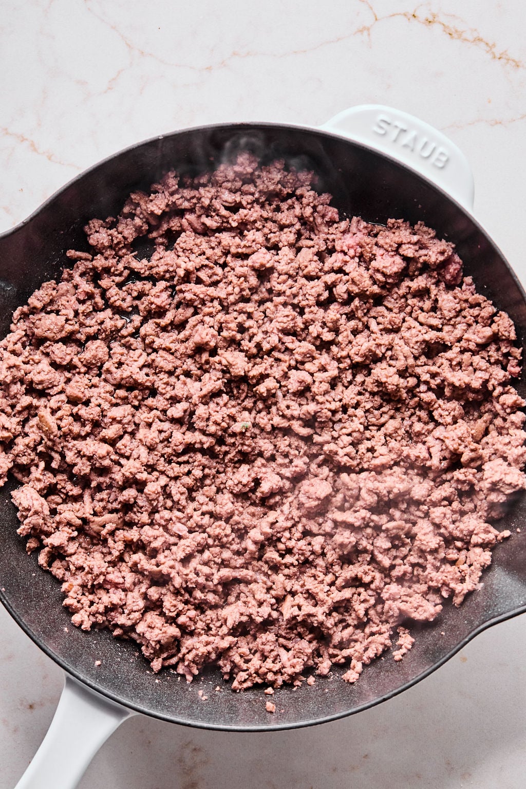 Cooked ground beef in a black skillet on a white marble countertop. Steam is visible rising from the meat.