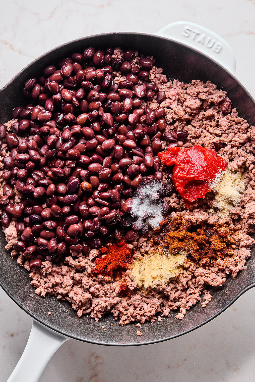 A skillet containing cooked ground beef, black beans, tomato paste, and various spices, all separated and not yet mixed together.