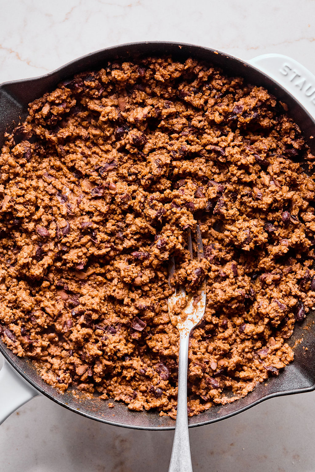 Cooked ground meat with spices in a black skillet, stirred with a fork, on a light-colored surface.