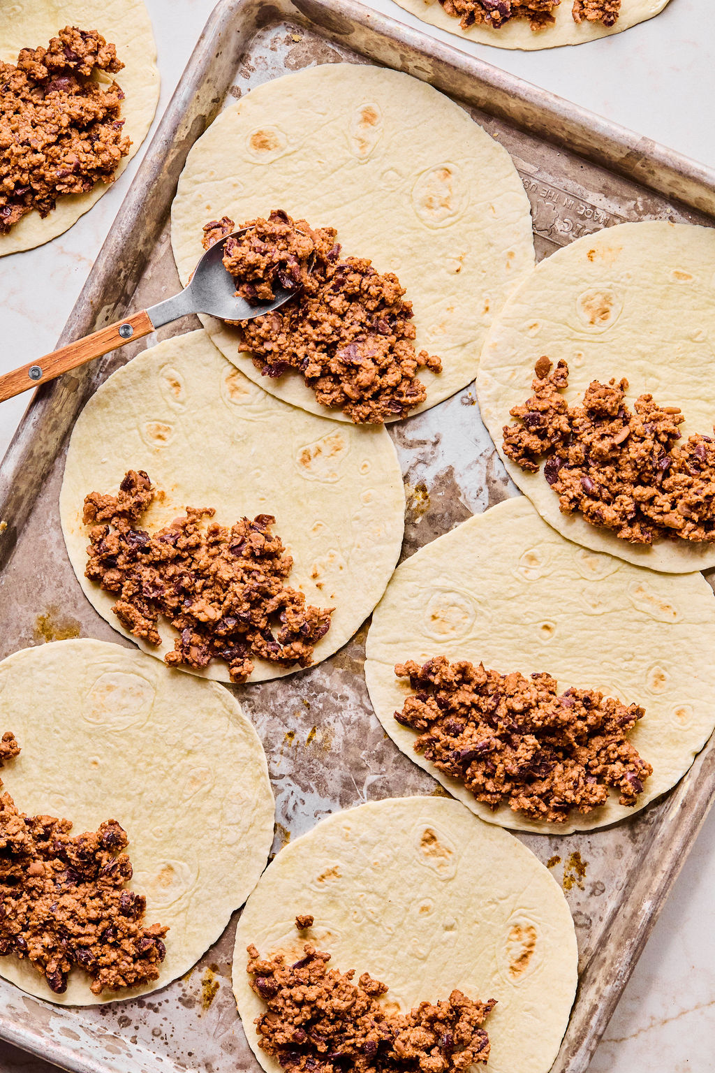 A baking sheet with several tortillas laid out, each topped with a portion of seasoned ground meat mixture, and a spoon resting on one tortilla.