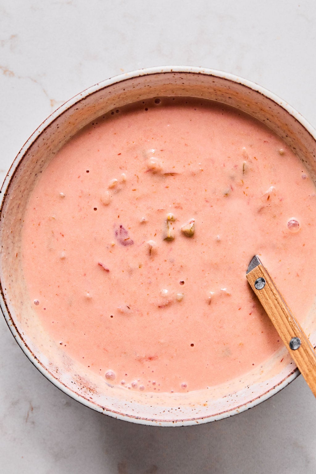 A bowl of creamy pink sauce with visible small vegetable pieces, stirred with a wooden-handled spoon, on a light countertop.