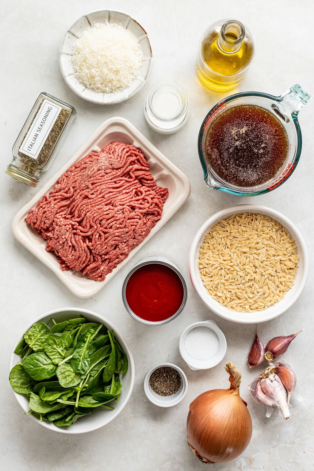 Ingredients for a recipe arranged on a white surface, including ground beef, orzo, spinach, onion, garlic, tomato sauce, broth, seasonings, oil, grated cheese, salt, and pepper.