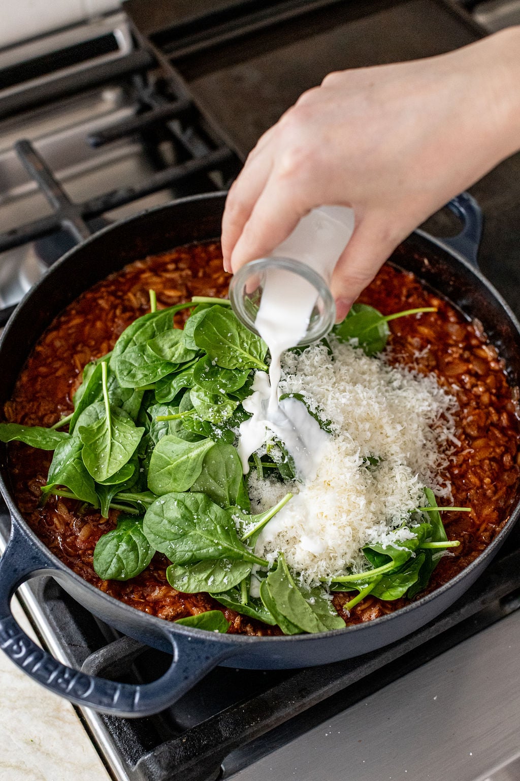 A hand pours cream into a pan of tomato sauce with fresh spinach and grated cheese on a stove.