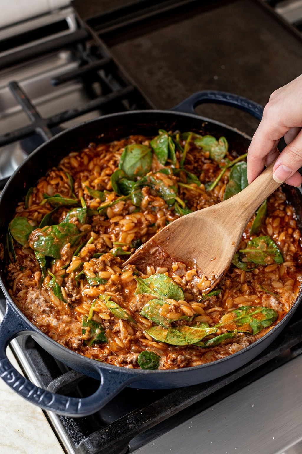 A hand stirs a skillet of cooked orzo pasta with spinach, tomato sauce, and cheese on a stovetop.