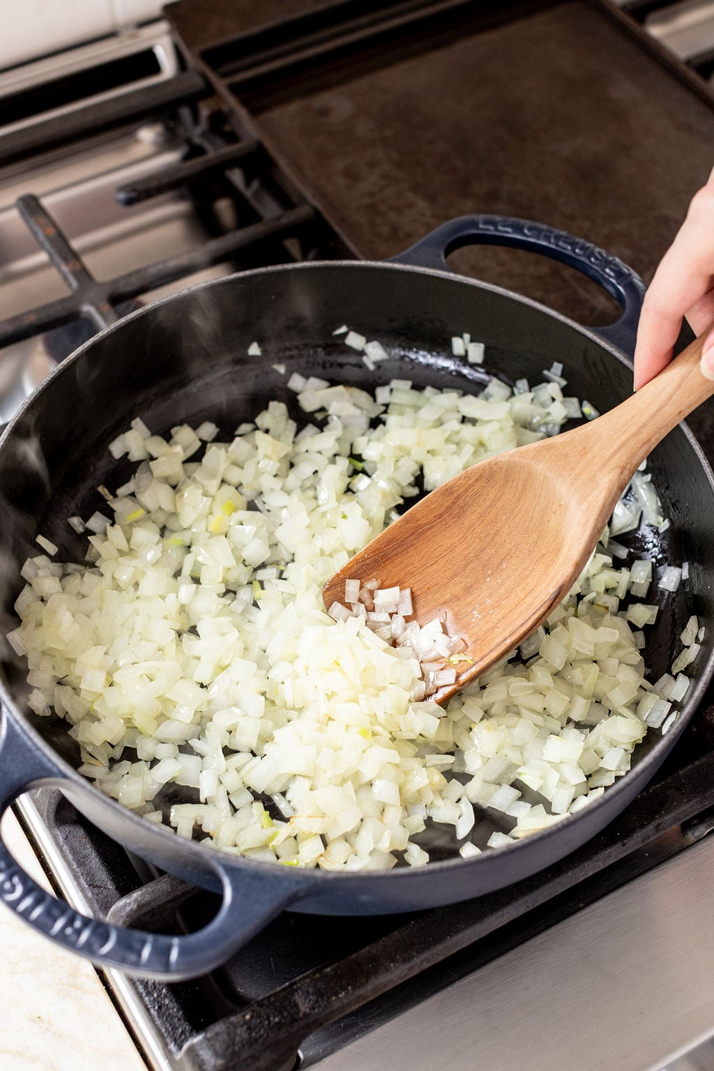Chopped onions being saut&eacute;ed in a black skillet on a stove, stirred with a wooden spatula.