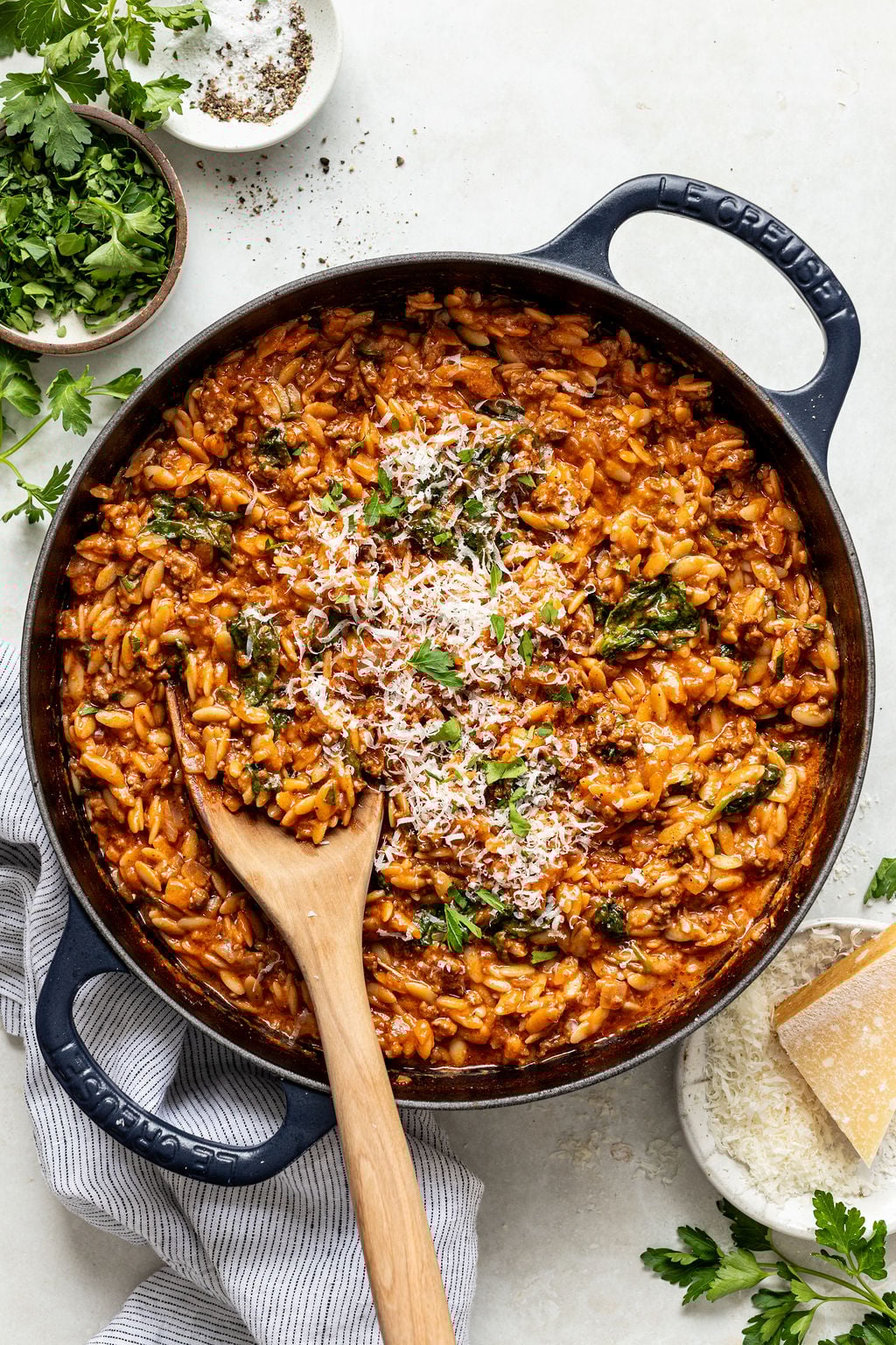 A skillet filled with tomato-based orzo pasta, topped with grated cheese and garnished with parsley, with a wooden spoon resting inside. Bowls of herbs and cheese sit nearby.