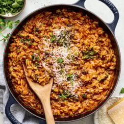 A skillet filled with tomato-based orzo pasta, topped with grated cheese and parsley, with a wooden spoon resting inside.