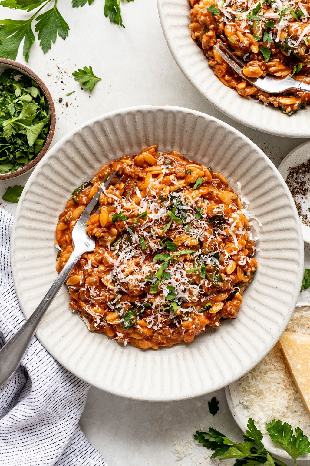A white bowl filled with creamy tomato-based orzo, topped with grated cheese and chopped parsley, with a fork resting on the side.