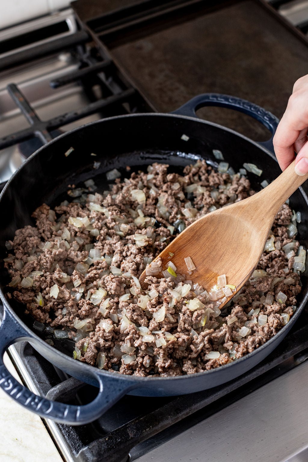 Ground beef and chopped onions are being cooked in a black skillet on a stovetop, with a wooden spoon stirring the mixture.