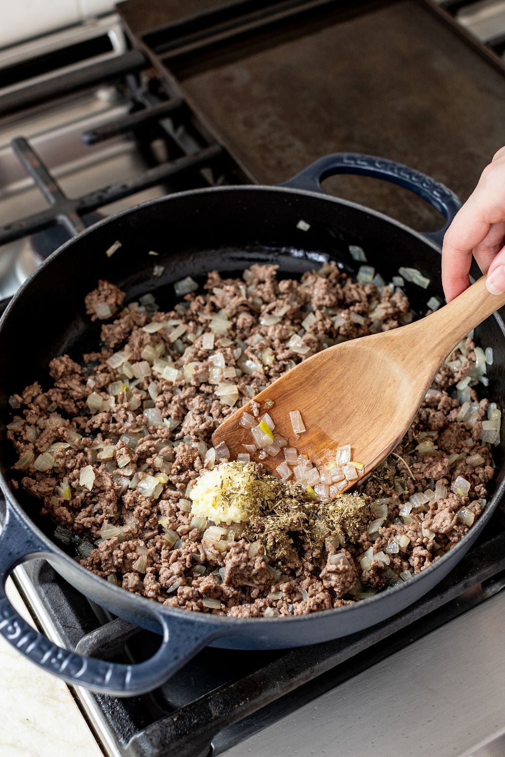 A hand stirs ground beef, chopped onions, herbs, and butter in a black skillet on a stovetop with a wooden spoon.