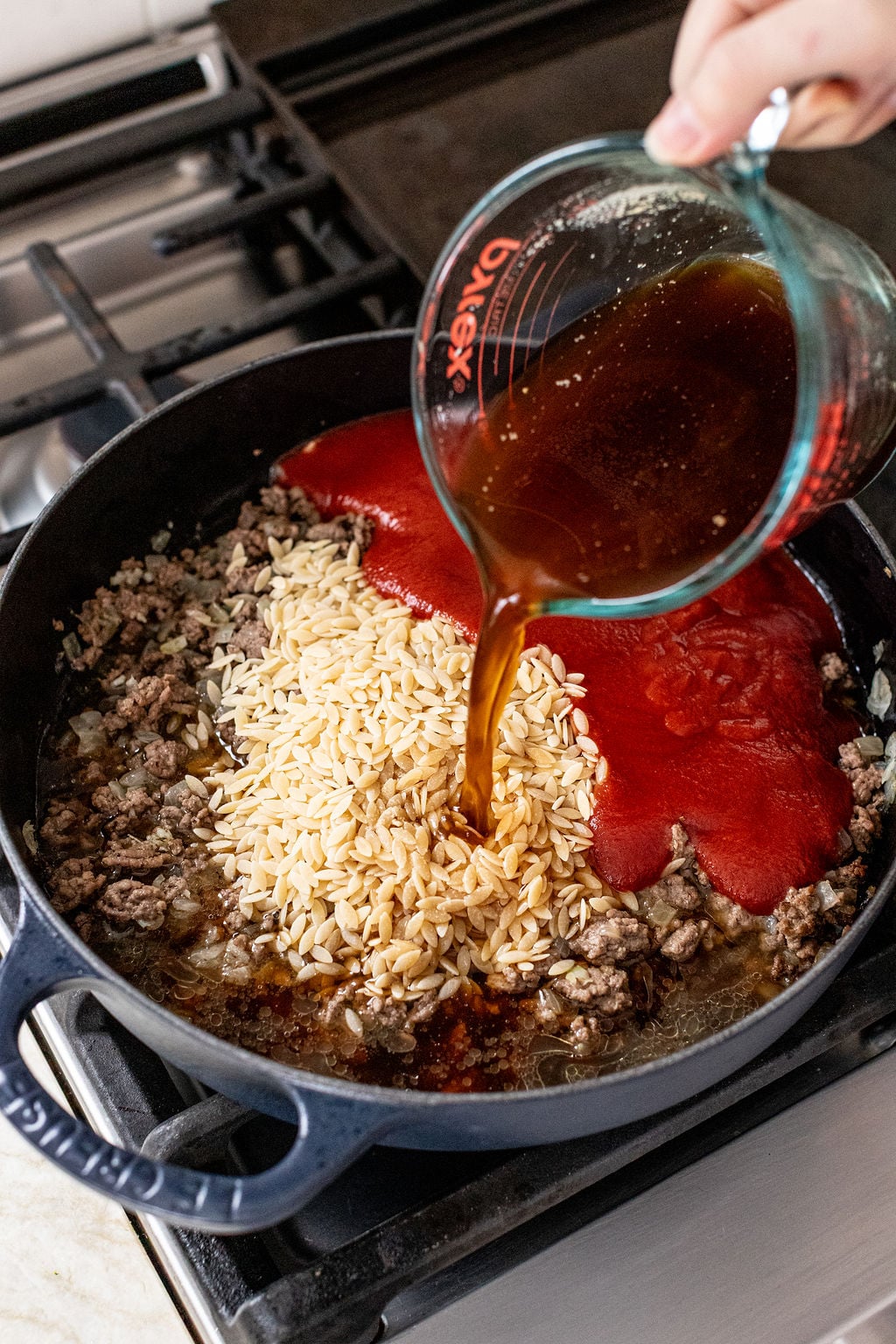 A hand pours broth from a measuring cup into a skillet containing ground meat, dry orzo, and tomato sauce on a stovetop.