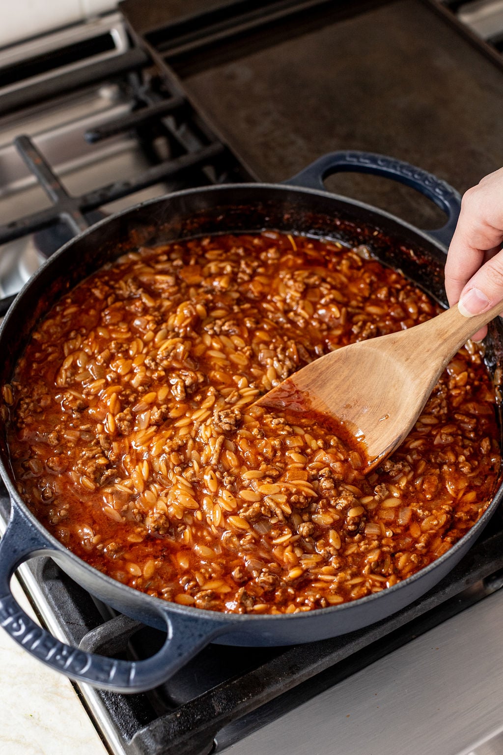 A hand stirs a pot of orzo pasta in tomato sauce with a wooden spatula on a stovetop.