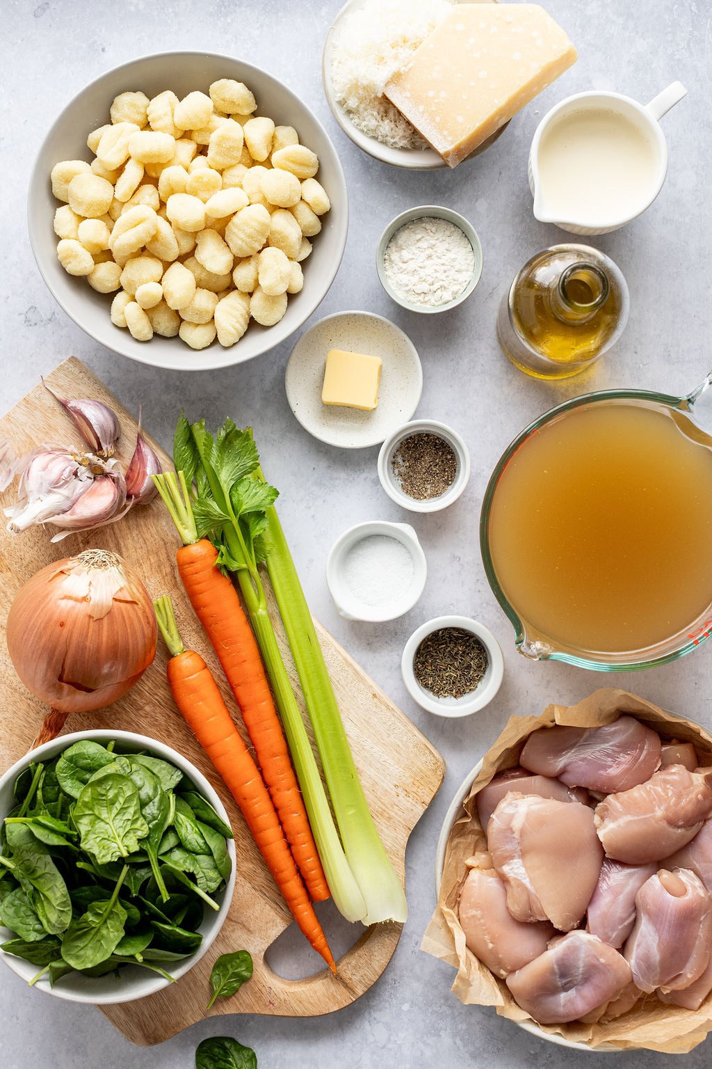 Top-down view of ingredients for a soup, including chicken, gnocchi, broth, carrots, celery, onion, spinach, cheese, cream, olive oil, butter, garlic, and seasonings.