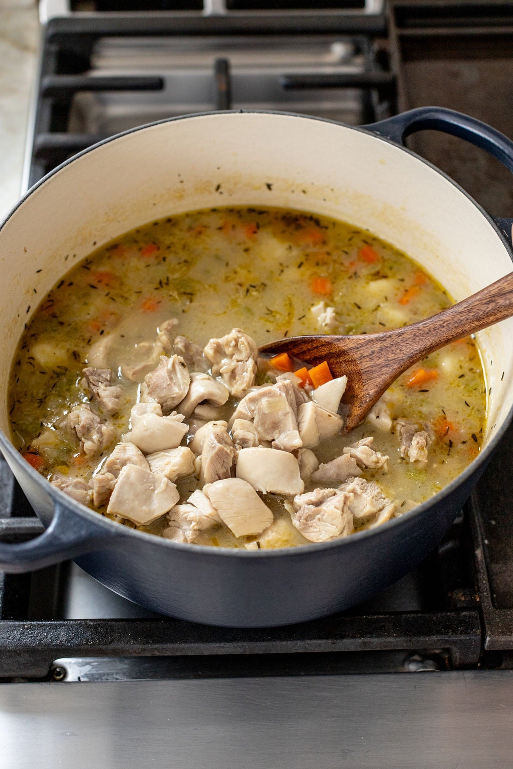 A pot of soup with diced chicken, carrots, and herbs being stirred with a wooden spoon on a stovetop.