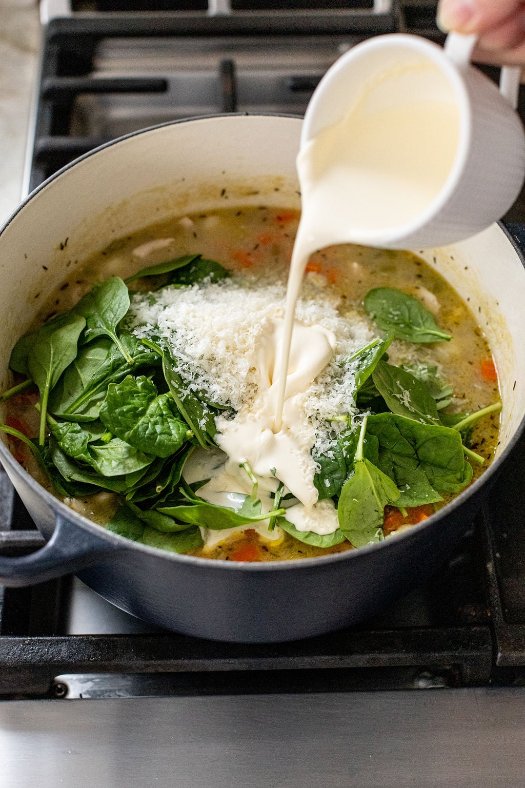 Cream is being poured into a pot of soup containing fresh spinach, grated cheese, and chopped vegetables on a stove.