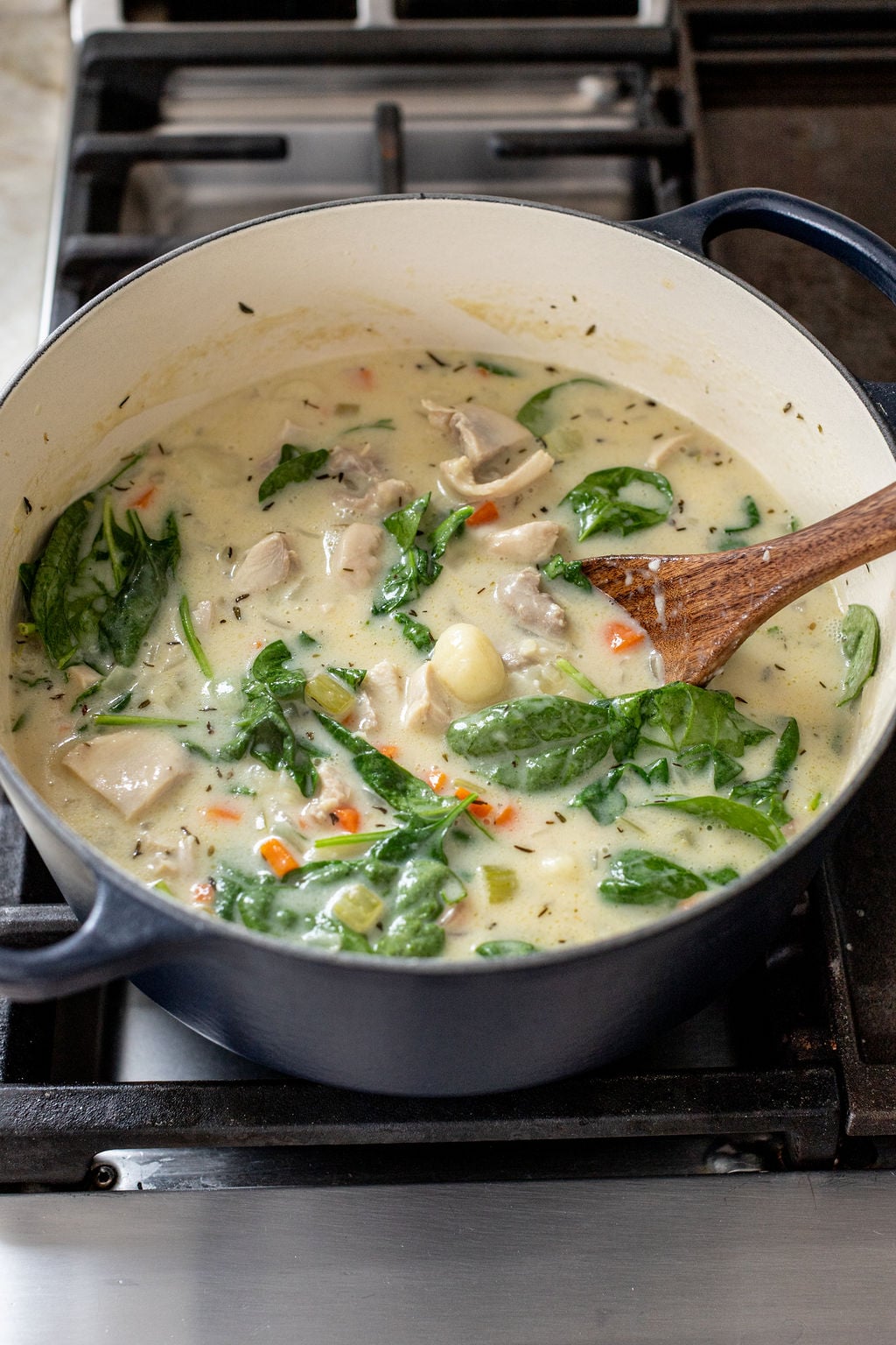 A pot of creamy chicken and vegetable soup with spinach sits on a stove, being stirred with a wooden spoon.