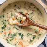 A pot of creamy soup with gnocchi, diced carrots, spinach, herbs, and chunks of chicken is being stirred with a wooden spoon.