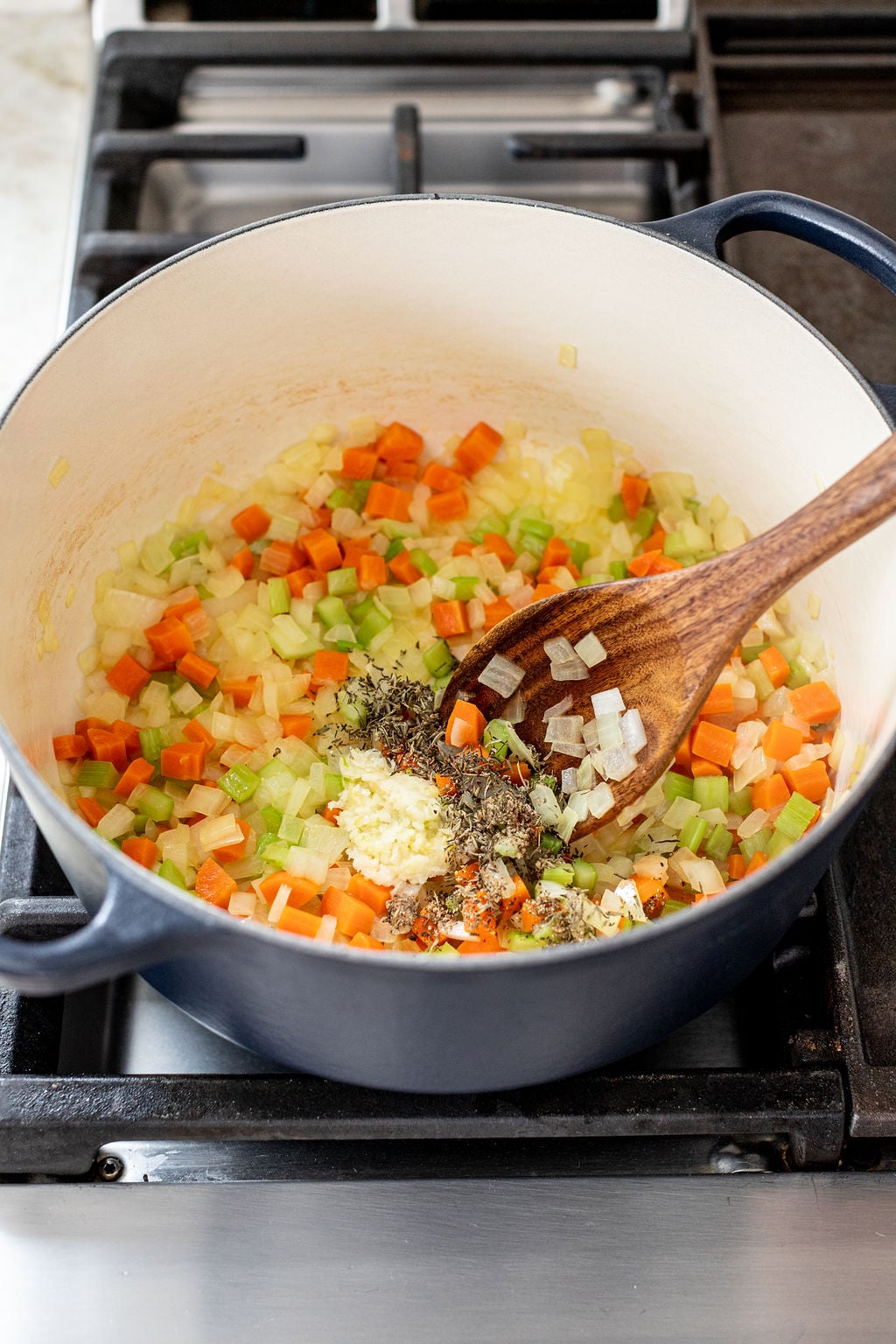 A pot on a stove contains chopped carrots, celery, onions, minced garlic, and herbs being stirred with a wooden spoon.