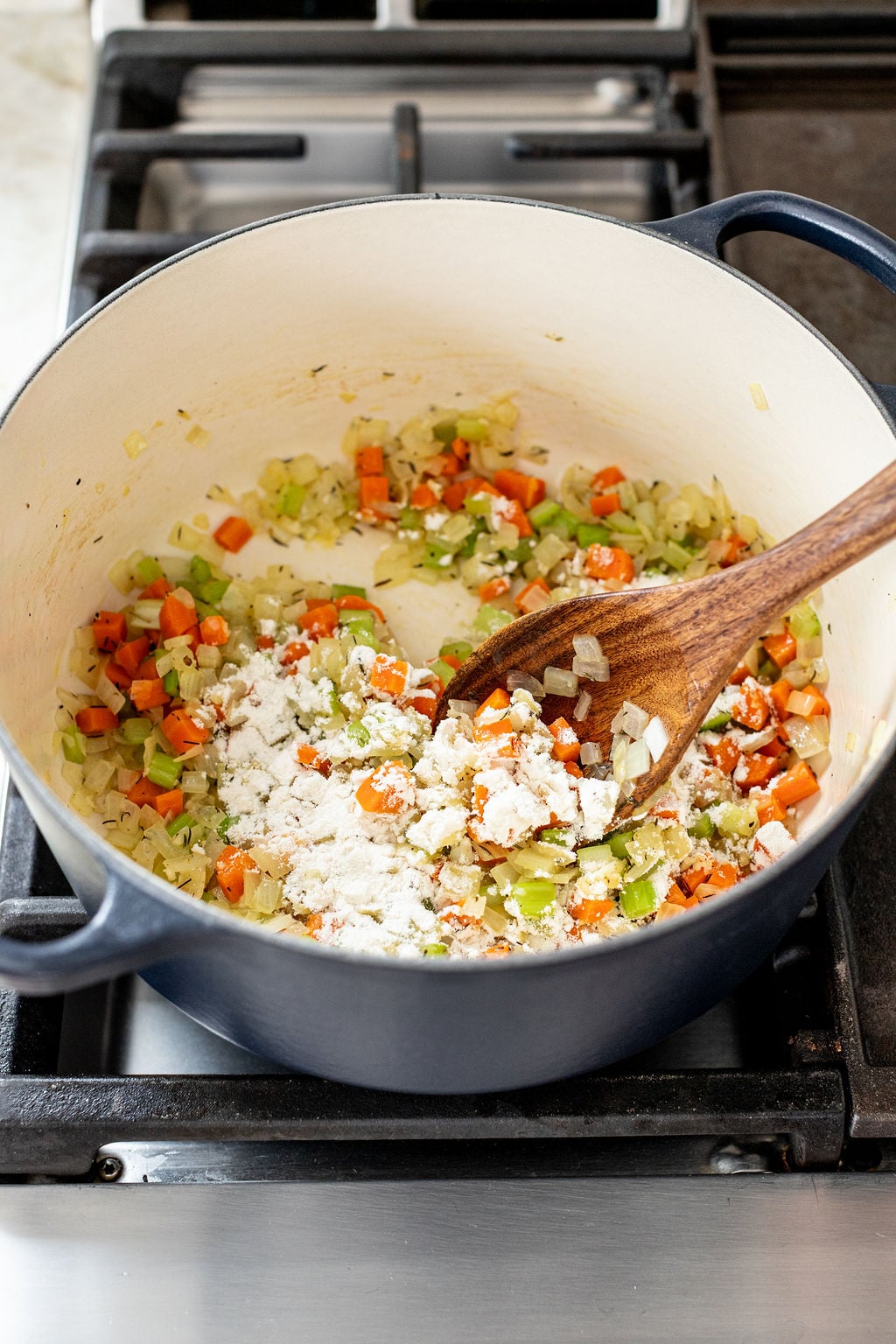 A pot on a stovetop contains chopped carrots, celery, onions, and flour being stirred with a wooden spoon.