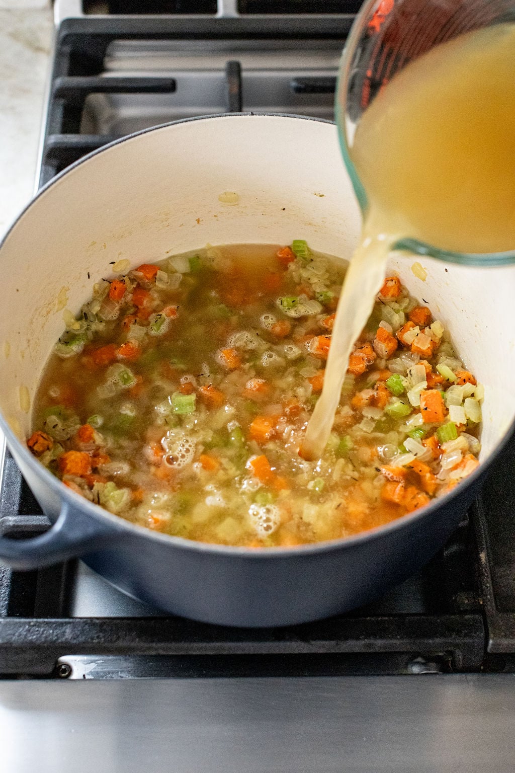 Vegetable broth is being poured into a pot containing diced carrots, celery, and onions on a stovetop.