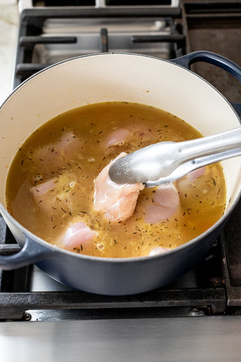 Raw chicken pieces being placed into a pot of seasoned broth with metal tongs on a stove.