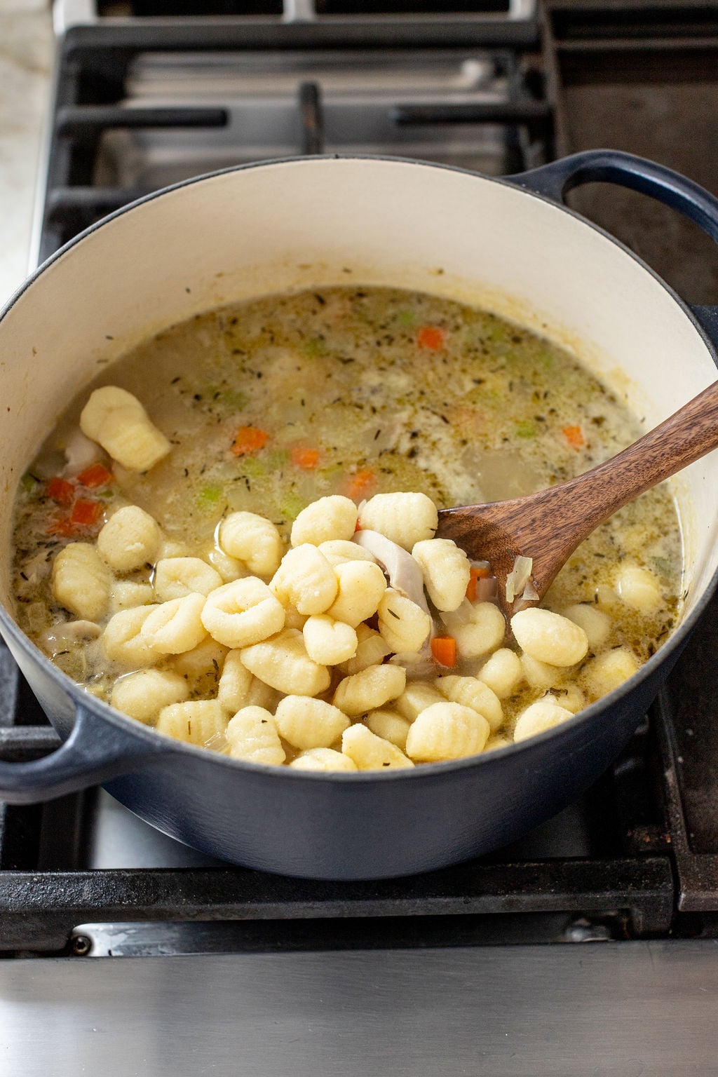 A pot of soup with gnocchi being stirred in with a wooden spoon on a stovetop.