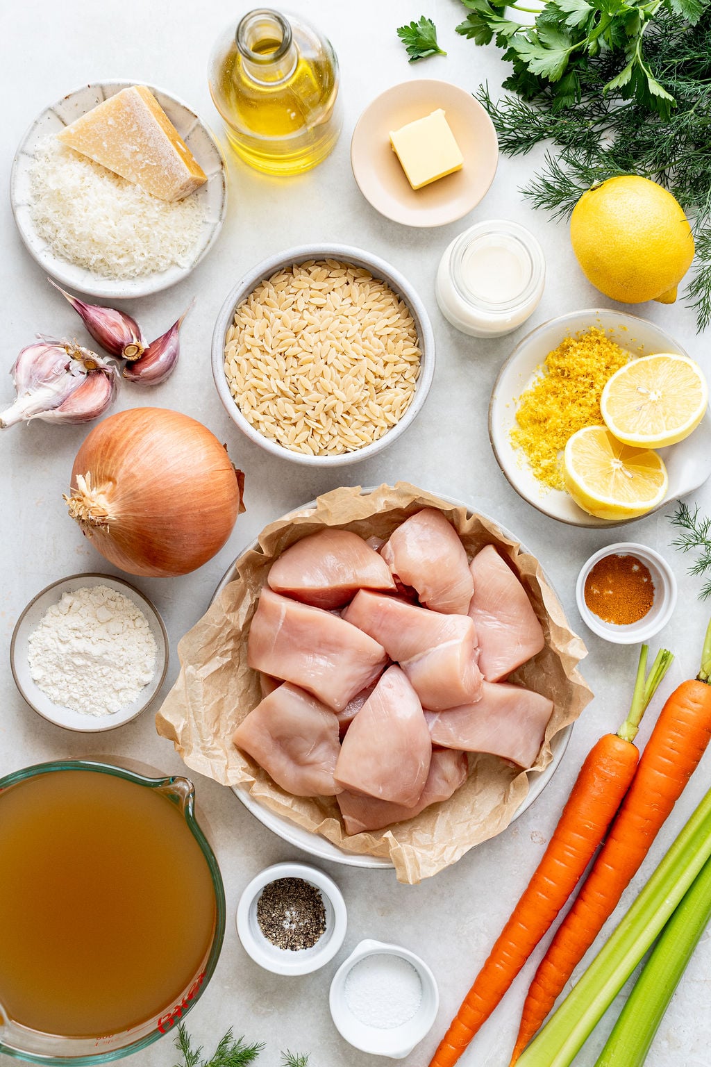 Raw chicken, orzo, carrots, onion, garlic, lemon, broth, cheese, cream, butter, herbs, flour, oil, salt, and pepper on a table.