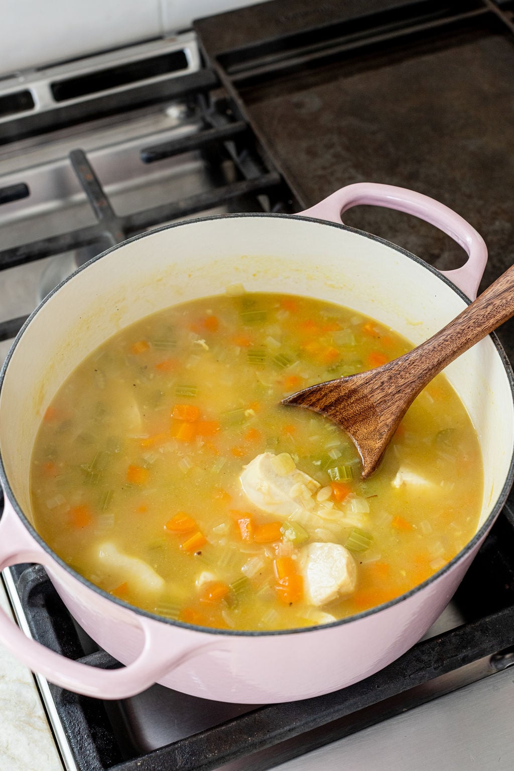 A pot filled with simmering chicken soup with carrots, celery, and broth sits on a stovetop with a wooden spoon inside.