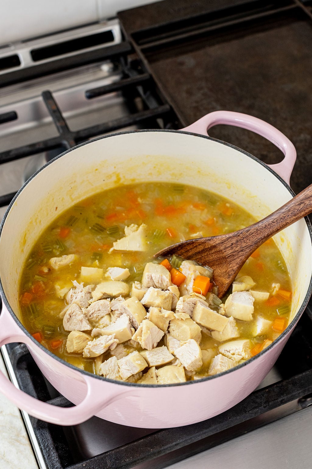 A pot on a stove contains soup with diced chicken, carrots, and potatoes; a wooden spoon rests inside the pot.