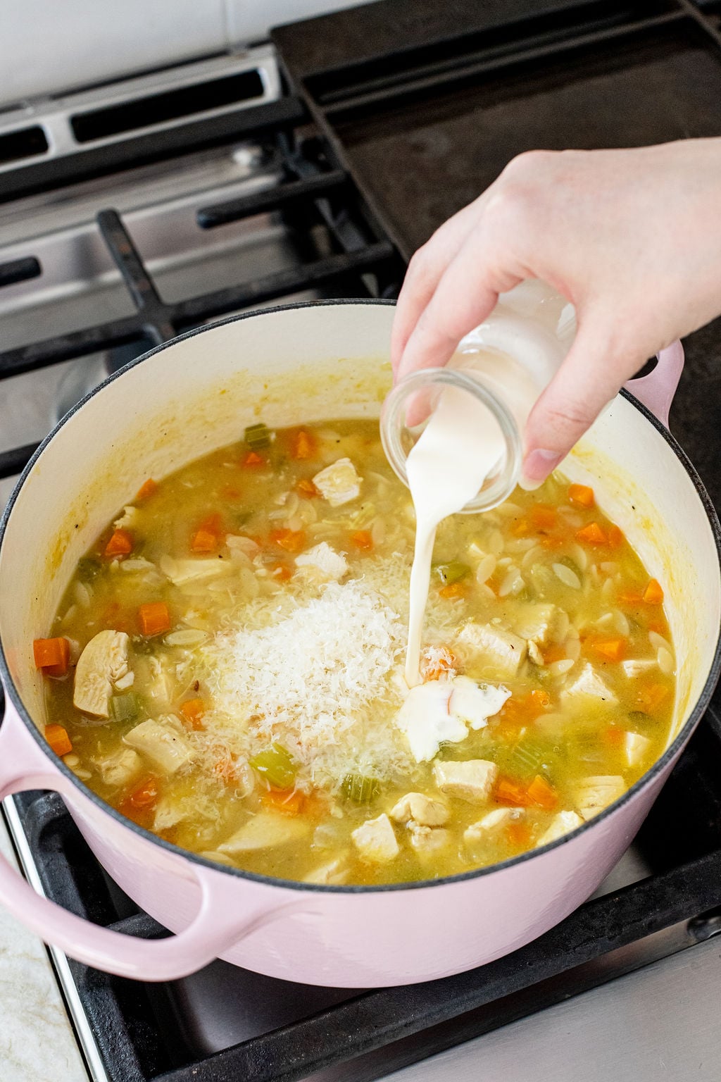 A hand pours cream into a pot of soup with diced chicken, carrots, celery, and grated cheese on a stove.