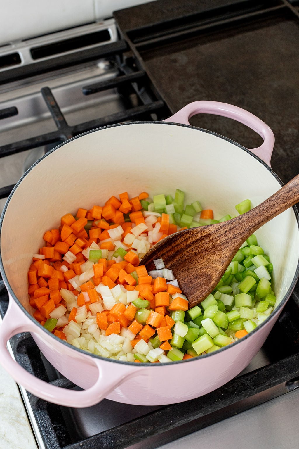 Chopped carrots, celery, and onions are being saut&eacute;ed in a pink Dutch oven on a stovetop, stirred with a wooden spoon.