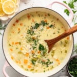 A pot of creamy soup with chunks of vegetables and herbs, being stirred with a wooden spoon, surrounded by lemon slices, cheese, and fresh herbs on a white surface.