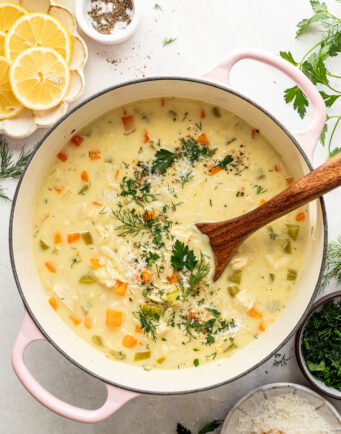 A pot of creamy soup with chunks of vegetables and herbs, being stirred with a wooden spoon, surrounded by lemon slices, cheese, and fresh herbs on a white surface.
